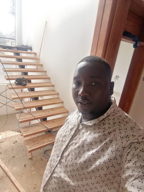 This staircase design in Kiambu allows natural light from the large windows to flow through the center of the house rather than being blocked by a heavy concrete staircase. The black ironmongery provides a modern, industrial contrast to the warmth of the mahogany frames.